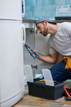 Professional repairman looking focused, using pipe wrench while examining and fixing sink pipe in the kitchenの写真素材
