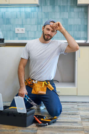 Quality work. Attractive young handyman, plumber wearing tool belt smiling at camera while holding a pipe wrench, ready for fixing sink pipe in the kitchenの写真素材