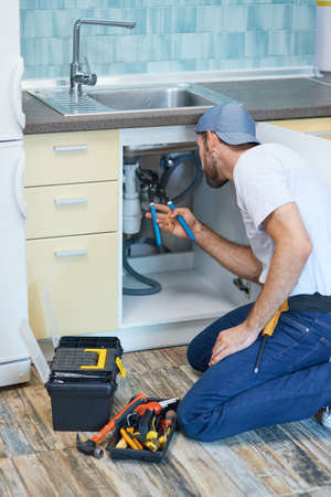 Plumbing troubles. Professional plumber using pipe wrench while examining and fixing sink pipe in the kitchenの写真素材