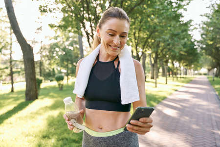 Energize your morning. Cheerful fit woman in sportswear refreshing, drinking water and using her smartphone after jogging in a green park on a sunny dayの写真素材