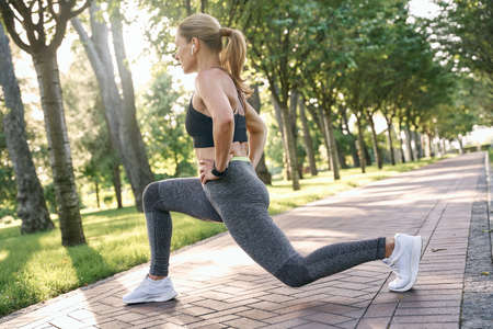 Feel fit. Sporty middle aged woman in sportswear looking focused, stretching her legs while having workout in a green park on a sunny dayの写真素材