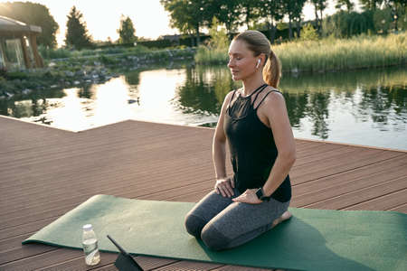 Replenish. Relaxed middle aged woman in sportswear smiling, sitting on a mat while doing yoga in nature on a sunny dayの写真素材
