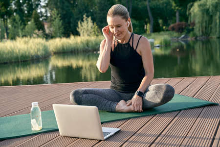 Sporty beautiful woman looking cheerful, watching tutorial on a laptop while doing yoga on a mat in nature on a sunny day outdoorsの写真素材