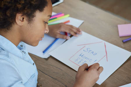 Portrait of mixed race teen schoolgirl drawing Stay Home inscription on paper with colorful markers...