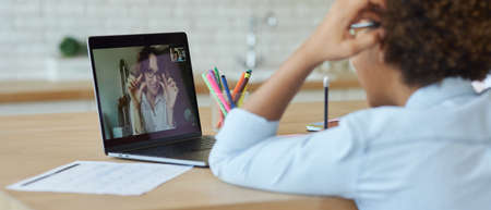 Rear view of mixed race teen schoolgirl drawing on paper with marker, using laptop while having online video lesson with teacher, sitting at home