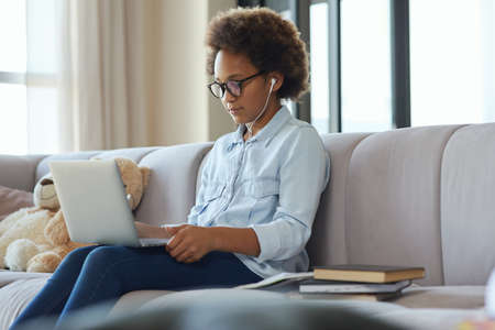 Mixed race teen girl wearing earphones using laptop, watching video while sitting on the sofa at...
