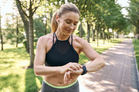 Morning jog. Beautiful sporty woman in sportswear and earphones looking at her watch, checking the result while running in a green park on a sunny summer dayの写真素材