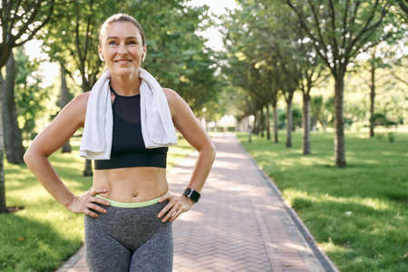 Stay active. Happy sporty woman in sportswear and wireless earphones smiling away, standing with towel around her neck after jogging in a green park on a sunny dayの写真素材