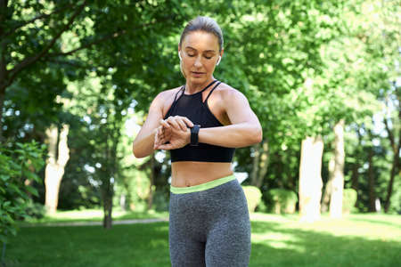 Jog in the morning. Beautiful sporty woman in sportswear and earphones looking at her watch and checking the result while jogging in a green park on a sunny summer dayの写真素材