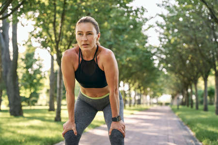 Rest for a while. Beautiful sporty woman in sportswear taking a break while jogging in a green park on a sunny summer dayの写真素材