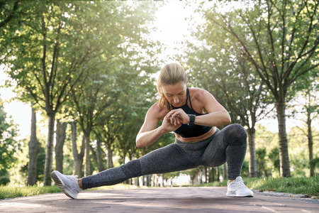 Endurance. Attractive middle aged athletic woman in sportswear looking focused, stretching her legs while training in a green park on a sunny dayの写真素材