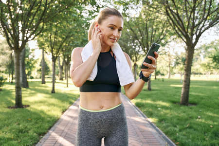 Use technology. Cheerful fit woman in sportswear and wireless earphones using her smartphone while jogging in a green park on a sunny dayの写真素材