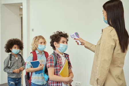 Cheerful boy wearing face mask watching his teacher measuring temperature, screening kid with digital thermometer while students coming to schoolの写真素材