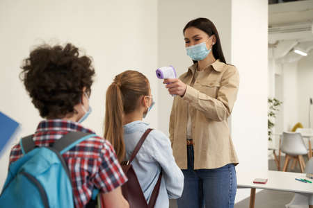 Nice young female teacher wearing protective mask smiling while screening school children using digital thermometer while kids coming to schoolの写真素材