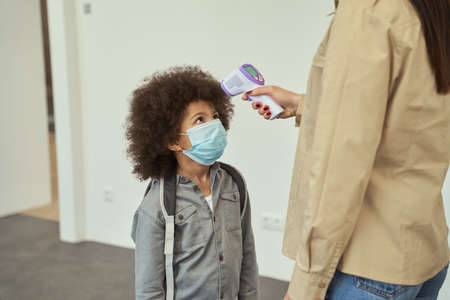 Cute little school boy wearing face mask watching his teacher measuring temperature, screening kid with digital thermometerの写真素材