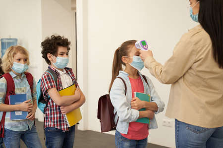 Queue. Young female teacher wearing protective mask measuring temperature, screening kids with digital thermometer while students coming to schoolの写真素材