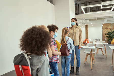 Young female teacher screening school children for fever against the spread of Covid19, using digital thermometer while kids coming to schoolの写真素材