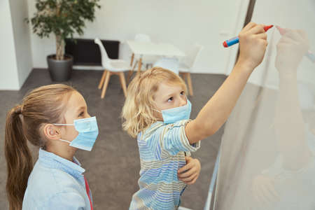 Little school kids, boy and girl wearing protective mask during coronavirus pandemic. Boy writing on board in elementary school classroomの写真素材