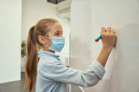 Portrait of smart little schoolgirl wearing protective mask during coronavirus pandemic writing numbers on board in elementary school classroomの写真素材