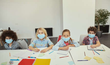 Group of diverse elementary school kids wearing protective mask during coronavirus pandemic looking cheerful while sitting together at the table in a classroomの写真素材