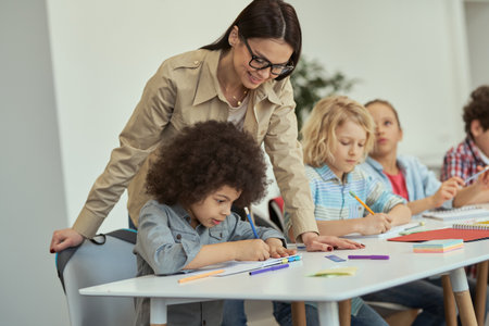 Caring young female teacher in glasses helping little schoolboy. Kids sitting at the table, studying in elementary school classroomの写真素材