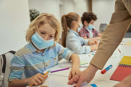 Little boy wearing protective face mask reading the task. Elementary school kids sitting together at the table in a classroomの写真素材