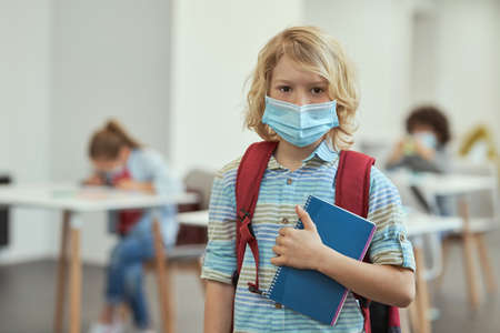 Portrait of calm little school boy wearing mask to prevent the spread of Covid19 looking at camera,...