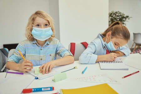 Little boy wearing protective face mask looking away while listening attentively. Elementary school kids sitting together at the table in a classroomの写真素材