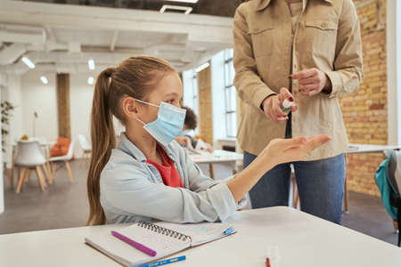 Hygiene first. Young female teacher using an alcohol spray to disinfect student hands in classroomの写真素材
