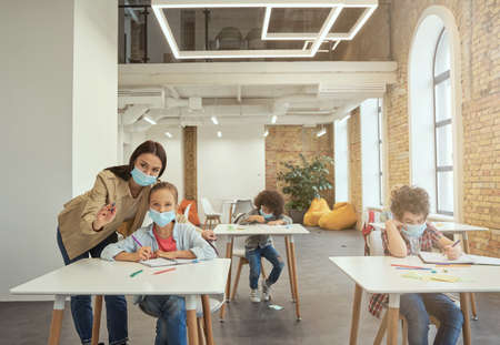 Attractive young female teacher wearing protective face mask helping little girl, explaining task. Kids studying in elementary school, sitting at the deskの写真素材