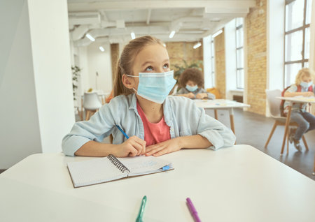 Gorgeous little girl wearing protective face mask during coronavirus outbreak looking away and listening attentively. Kid studying in elementary school, sitting at the deskの写真素材