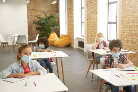 Wear face coverings indoors. School kids wearing protective face mask during coronavirus outbreak studying in elementary school, sitting at the desk in classroomの写真素材