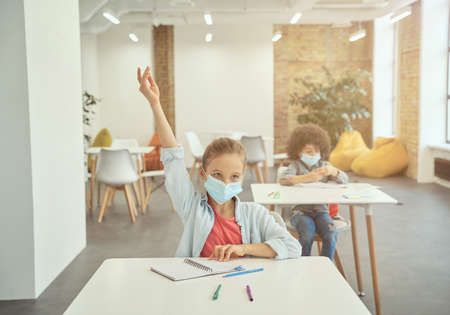 Little girl wearing protective face mask during coronavirus outbreak raised her hand in class. School child studying in elementary school, sitting at the deskの写真素材
