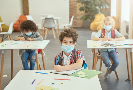 Little school boy wearing face mask during coronavirus outbreak looking at camera while sitting at the desk in classroom. Kids studying in elementary schoolの写真素材