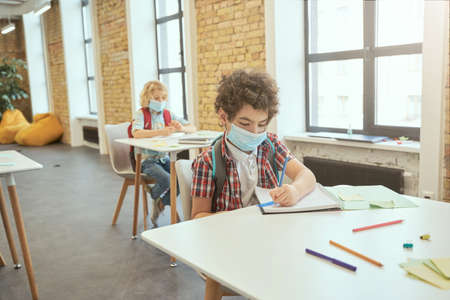 School boy wearing face mask during corona virus and flu outbreak writing in his notebook while sitting at the desk in a classroom. Kids studying in elementary schoolの写真素材