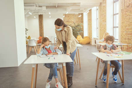 Full length shot of female teacher wearing protective face mask during coronavirus outbreak helping little girl in classroom. School kid studying in elementary school, sitting at the deskの写真素材