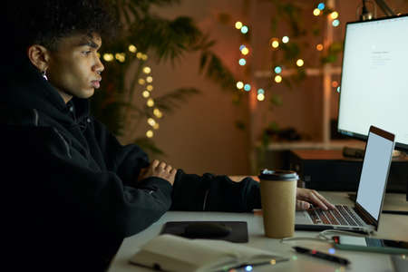 Side view of stylish young guy looking at monitor screen while working on laptop computer at late nightの写真素材