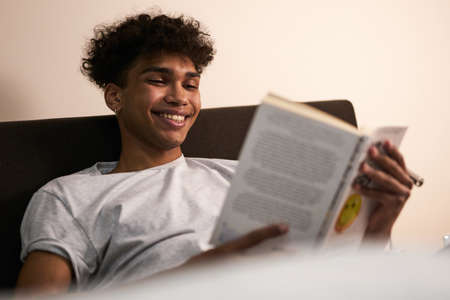 Amazing story. Portrait of cheerful brunette guy lying in the bed at home, smiling while reading interesting book at late eveningの写真素材