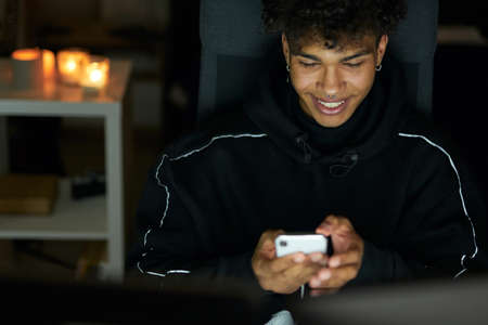 Social media. Smiling young handsome guy looking happy while texting, chatting with friends, using smartphone late at night at homeの写真素材