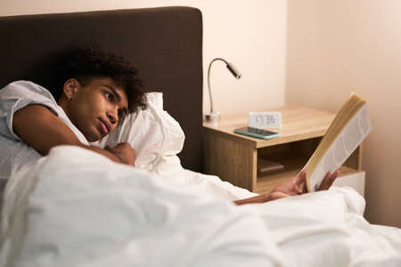 Young guy lying in the bed at home, resting and reading an interesting book before going to sleepの写真素材