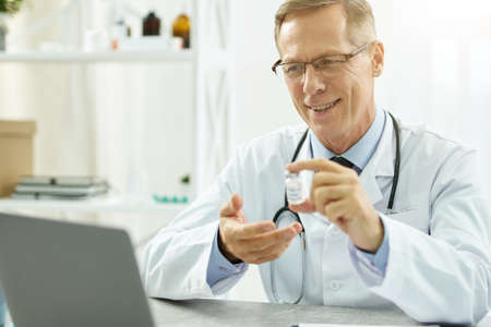 Joyful doctor holding covid-19 vaccine and using laptop in clinicの写真素材