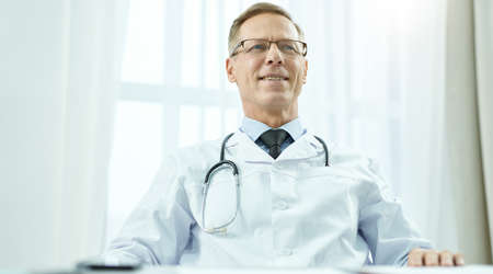 Cheerful male doctor sitting at the table in his officeの写真素材