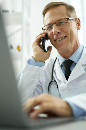 Cheerful doctor talking on mobile phone and using laptop in clinicの写真素材