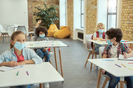Little boy wearing protective face mask during coronavirus outbreak raised his hand in class. School kids studying in elementary school, sitting at the deskの写真素材