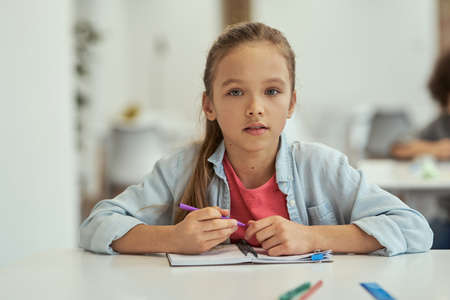 Sweet little school girl looking at camera while sitting at the desk in classroom