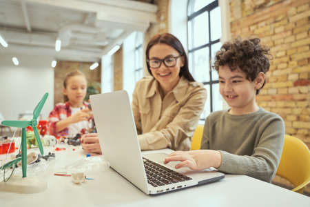 Young female teacher in glasses using laptop in classroom, showing scientific robotics video to little boyの写真素材