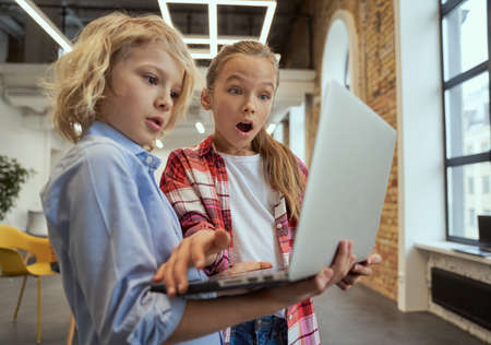 Two curious kids, little boy and girl looking surprised while using laptop, standing in a classroom during STEM lessonの写真素材