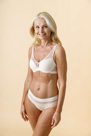 Studio shot of gorgeous mature woman with blonde hair in white underwear smiling while posing for camera, standing isolated over beige backgroundの写真素材