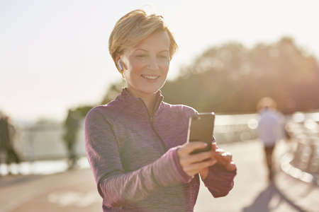 Results speak for themselves. Portrait of joyful active mature woman in sportswear using her smartphone, checking result after running outdoors on a sunny dayの写真素材