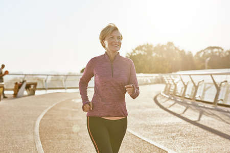 Improving health. Portrait of happy motivated mature woman in sportswear running outdoors on a sunny dayの写真素材